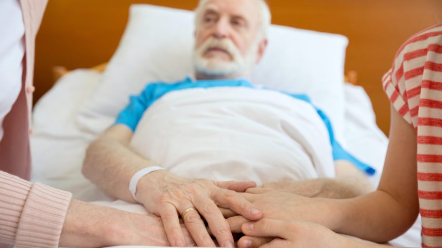 grandmother and grandfather holding hands with patient lying in hospital bed in clinic
