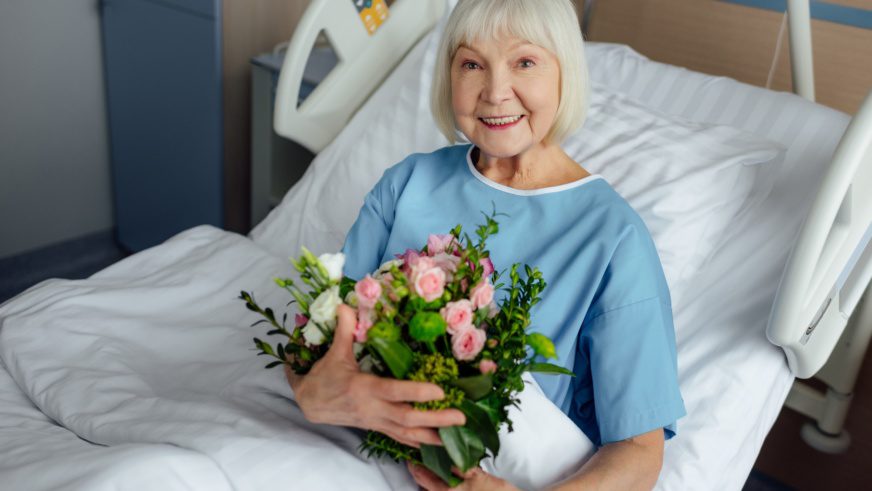 happy recovering senior woman lying in bed with flowers and looking at camera in hospital