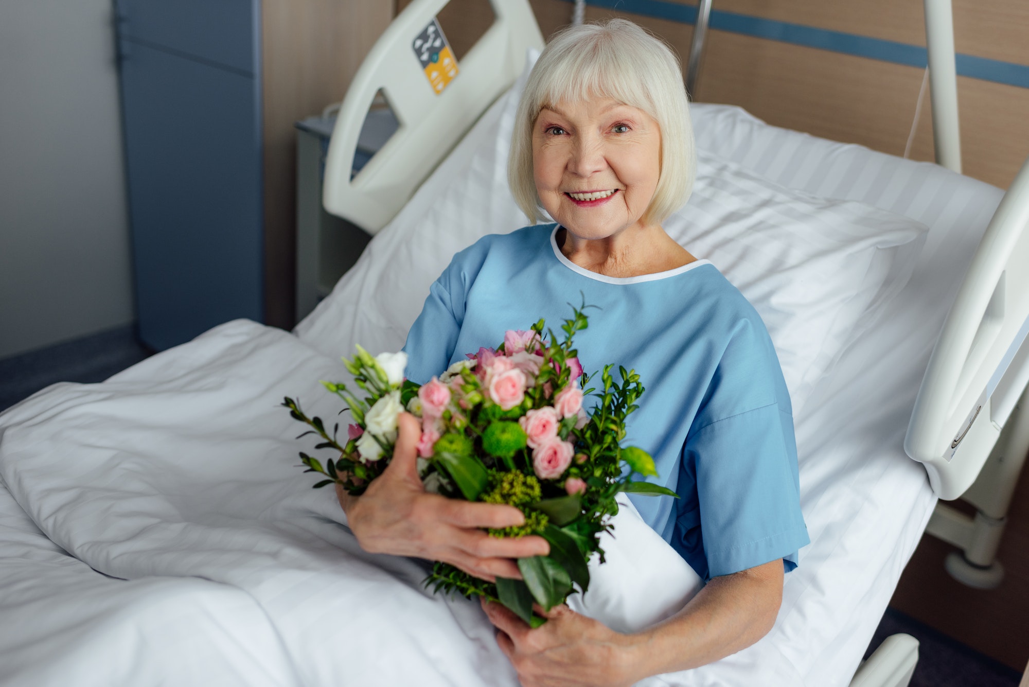 happy recovering senior woman lying in bed with flowers and looking at camera in hospital