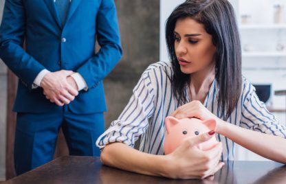 Worried woman covering piggy bank with hands at table near collector with clenched hands in room