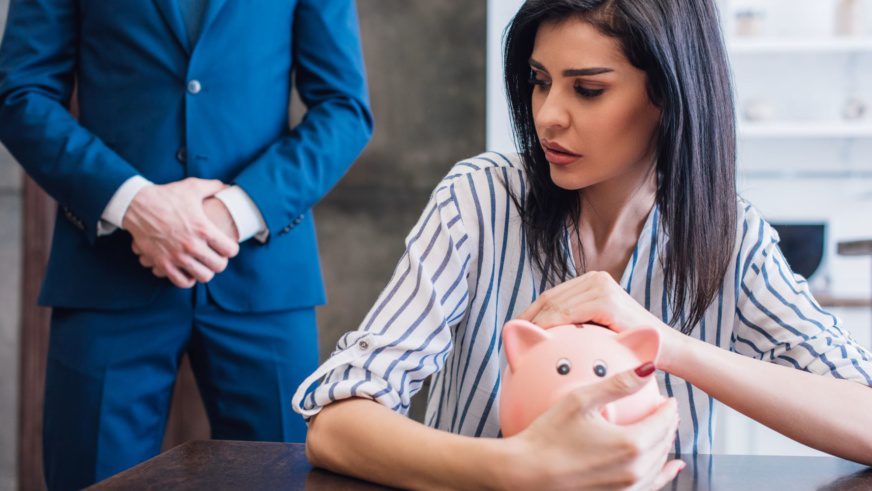 Worried woman covering piggy bank with hands at table near collector with clenched hands in room