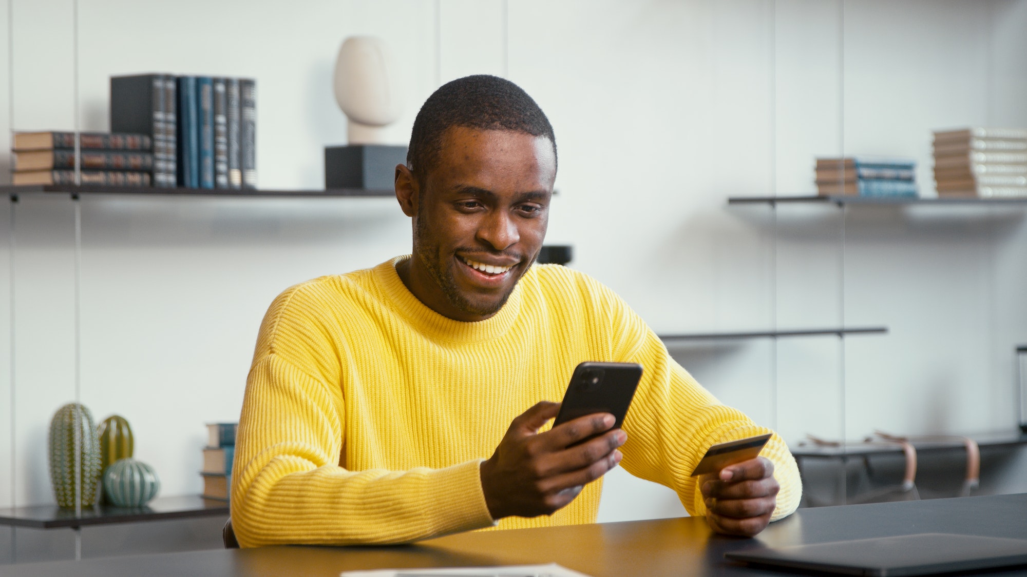 Smiling man in yellow shirt looks at bank card in hand and enters digits to smartphone sitting