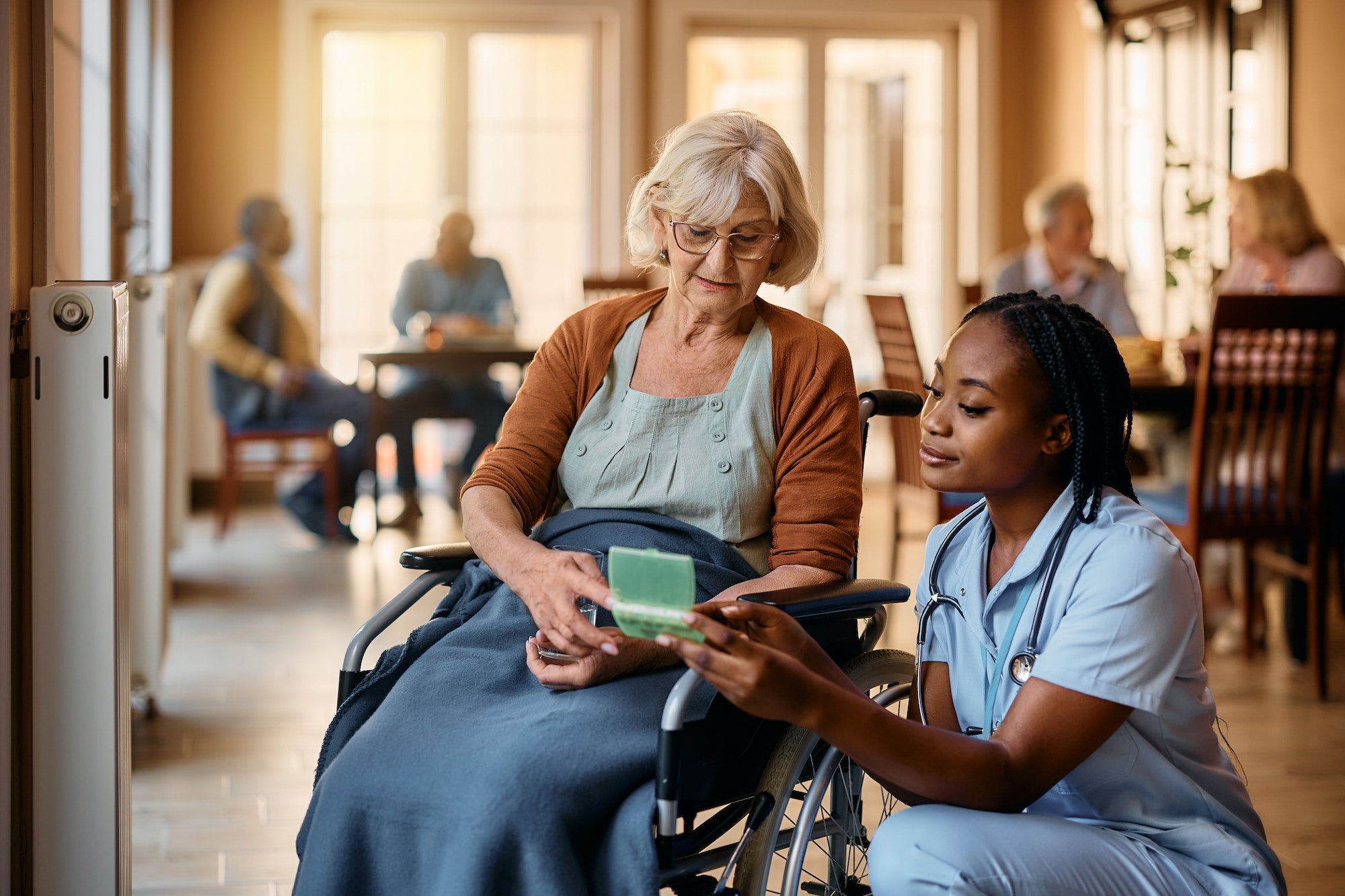 Young black nurse giving pills to senior woman in wheelchair at nursing home.